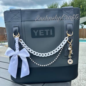 Può includere: Una borsa tote YETI grigio scuro con fiocco bianco, catena e ciondoli. La borsa ha una base nera e il testo "SnshnshCrafts" è visibile. La borsa è appoggiata su un tavolo grigio.
