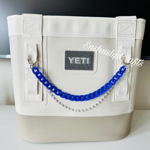 May include: A soft cooler bag with a two-tone design, featuring a light gray body and a darker gray base. It has white straps and a blue chain with a pearl strand. The YETI logo is visible on the front.