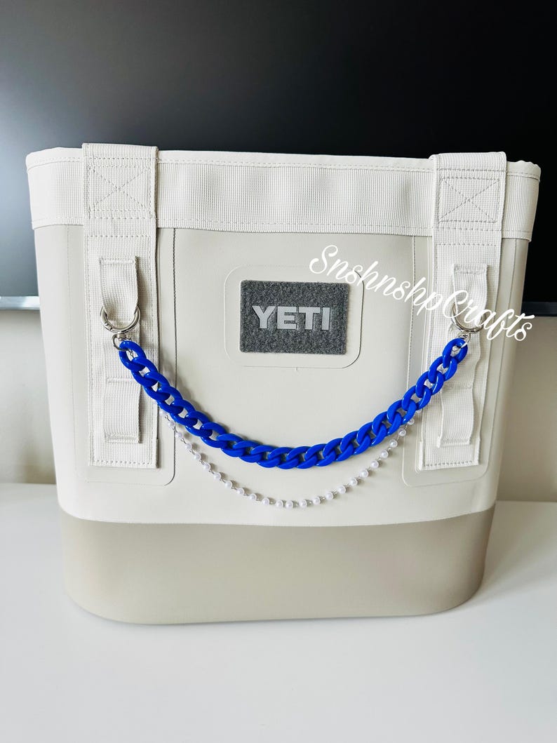 May include: A soft cooler bag with a two-tone design, featuring a light gray body and a darker gray base. It has white straps and a blue chain with a pearl strand. The YETI logo is visible on the front.