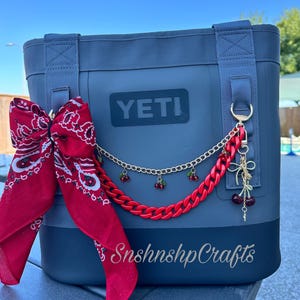 Op de afbeelding: Een marineblauwe YETI koeltas met een rode bandana strik en een decoratieve ketting. De ketting heeft rode plastic schakels, een gouden ketting en kersen bedels. De tekst "SnshnshpCrafts" is zichtbaar.