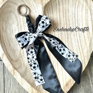 May include: A soccer-themed keychain featuring a black and white soccer ball ribbon bow and black satin ribbons. The keychain has a silver-colored ring and a braided black cord. The text "SnshnshpCrafts" is visible.