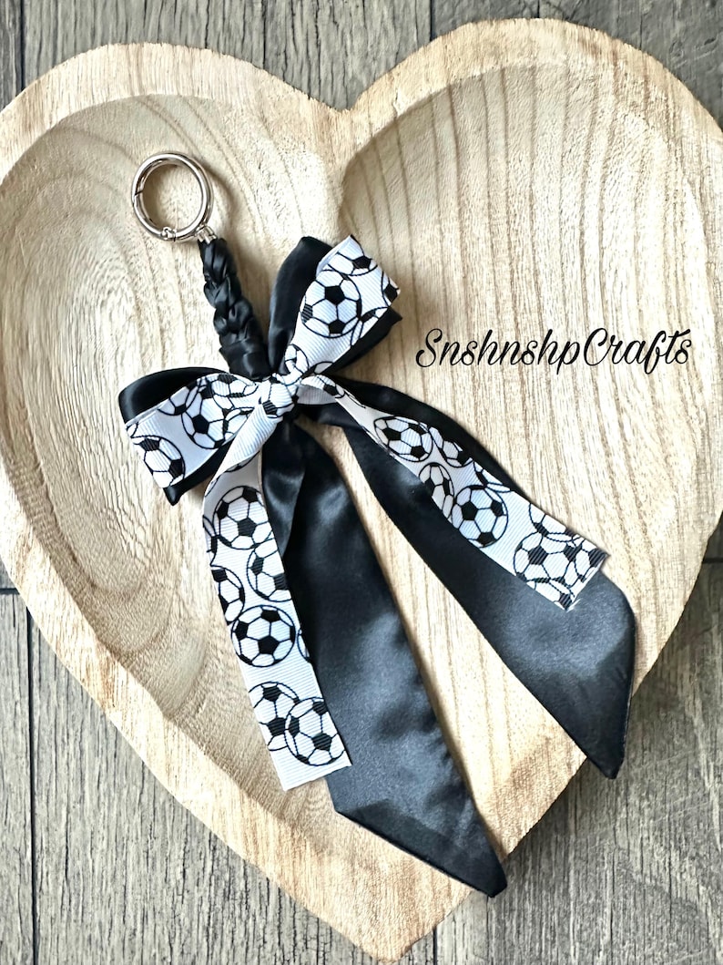 May include: A soccer-themed keychain featuring a black and white soccer ball ribbon bow and black satin ribbons. The keychain has a silver-colored ring and a braided black cord. The text "SnshnshpCrafts" is visible.
