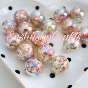 Puede incluir: Un conjunto de cuentas redondas y transparentes llenas de confeti colorido, dispuestas sobre un plato blanco en forma de coraz&oacute;n con lunares negros. Las cuentas tienen una calidad iridiscente. El texto "SnshnshpCrafts" es visible.