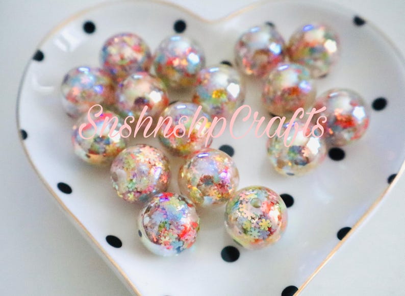 Puede incluir: Un conjunto de cuentas redondas y transparentes llenas de confeti colorido, dispuestas sobre un plato blanco en forma de coraz&oacute;n con lunares negros. Las cuentas tienen una calidad iridiscente. El texto "SnshnshpCrafts" es visible.