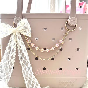 Puede incluir: Un bolso tote rosa claro con un lazo de encaje blanco y una cadena dorada con cuentas rosas. El bolso tiene un dise&ntilde;o perforado y el texto "SnshnshipCrafts" est&aacute; impreso en la parte delantera.
