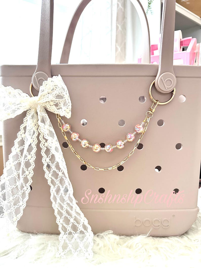 Puede incluir: Un bolso tote rosa claro con un lazo de encaje blanco y una cadena dorada con cuentas rosas. El bolso tiene un dise&ntilde;o perforado y el texto "SnshnshipCrafts" est&aacute; impreso en la parte delantera.