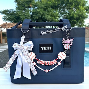 May include: A navy blue YETI tote bag featuring a white ribbon with a baseball pattern, a beaded accessory with baseball charms, and a custom name tag reading CONNER 11. A pink and white baseball MOM charm hangs from the right strap.