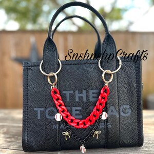 May include: Black tote bag with gold hardware and a red chain strap. The bag features the text "THE TOTE BAG" in large letters. The chain is adorned with baseball charms and bows. The bag is sitting on a wooden surface.