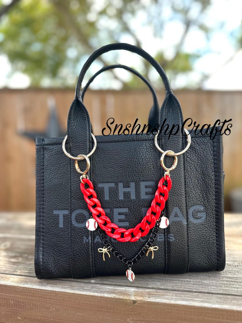 May include: Black tote bag with gold hardware and a red chain strap. The bag features the text "THE TOTE BAG" in large letters. The chain is adorned with baseball charms and bows. The bag is sitting on a wooden surface.