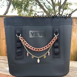 Puede incluir: Una bolsa nevera negra Yeti con una cadena decorativa. La cadena es de color cobre y tiene dijes, incluyendo balones de fútbol y un colgante. La bolsa tiene correas negras y la palabra "YETI" en la parte delantera.