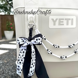 May include: A white and gray cooler with a soccer ball-themed accessory. The accessory includes a black and white soccer ball ribbon and a beaded strand with soccer ball accents. The cooler has the word "YETI" on it and the text "SnshnshpCrafts".
