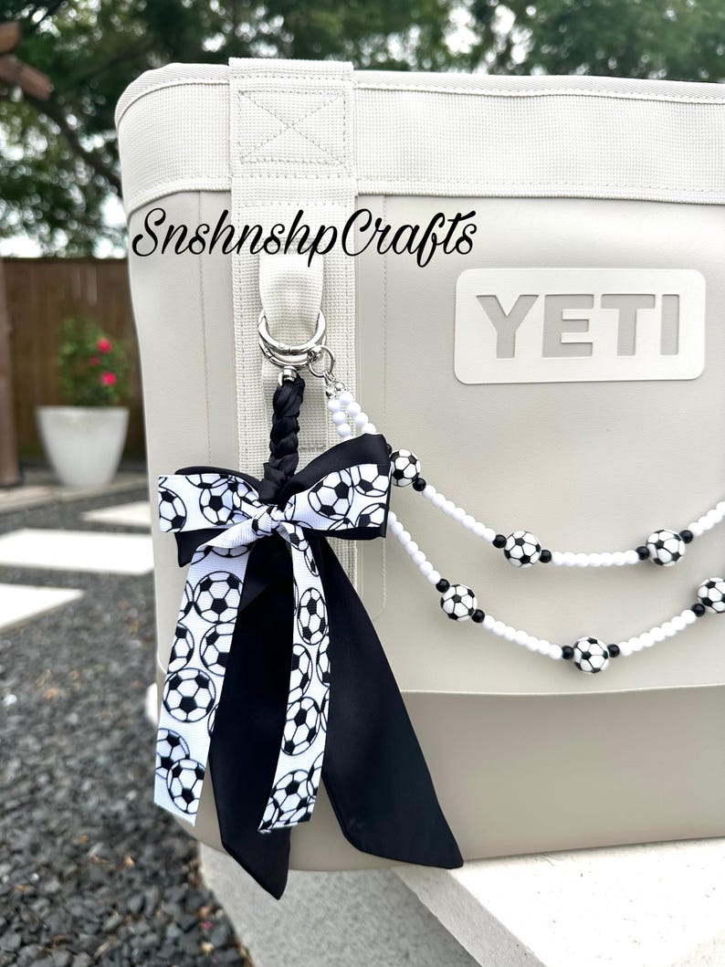 May include: A white and gray cooler with a soccer ball-themed accessory. The accessory includes a black and white soccer ball ribbon and a beaded strand with soccer ball accents. The cooler has the word "YETI" on it and the text "SnshnshpCrafts".