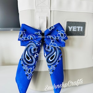 May include: A blue bandana bow attached to a white and gray cooler. The bow features a paisley design in white and black. The cooler has a gray Yeti logo and a white handle.