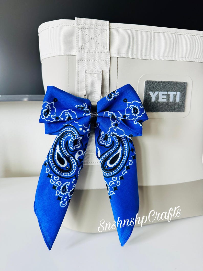 May include: A blue bandana bow attached to a white and gray cooler. The bow features a paisley design in white and black. The cooler has a gray Yeti logo and a white handle.