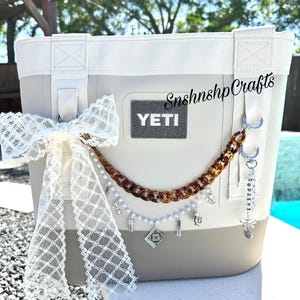 Pode incluir: Uma bolsa térmica YETI bege e branca com um laço de renda decorativo e uma corrente de berloques. A bolsa tem um patch cinza com o logotipo YETI e uma corrente marrom e creme com berloques. O texto "SnshnshpCrafts" é visível.