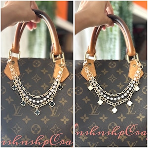 May include: A brown handbag with a gold chain and pearl accessory. The accessory features multiple gold chains, white pearls, and black clover charms. The bag has brown leather handles and gold hardware.