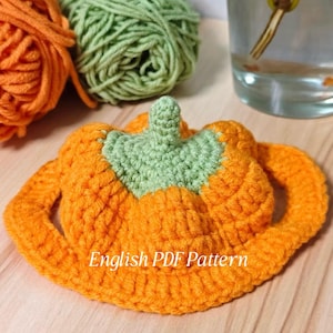 Op de afbeelding: Een gehaakte pompoen in oranje en groen met de tekst "English PDF Pattern". De pompoen heeft een groene steel en een ronde rand. Op de achtergrond zijn twee bollen wol te zien, één oranje en één groen.