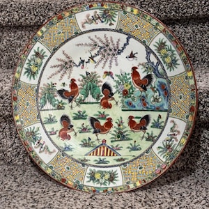 May include: A large, round decorative plate with a detailed rooster design. The plate features a central scene of roosters in a garden setting, surrounded by a patterned border with floral and geometric elements. The color palette includes red, green, yellow, and blue.
