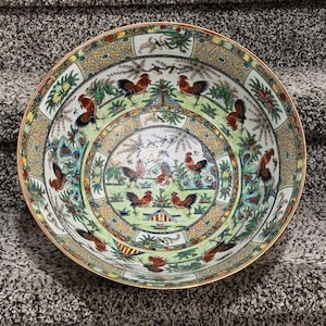 May include: A decorative ceramic bowl with a detailed rooster design. The bowl features a gold rim and a colorful pattern of roosters, foliage, and geometric shapes. The bowl is likely for decorative purposes.