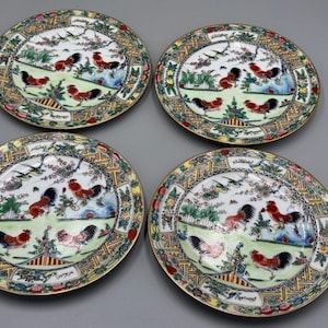 May include: Four decorative ceramic plates with a rooster motif. Each plate features a central scene with roosters, birds, and landscape elements, surrounded by a patterned border with floral and geometric designs. The plates have a gold rim.