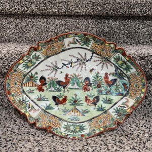 May include: An oval, antique porcelain serving platter with a scalloped edge. The platter features a hand-painted scene of roosters and foliage in shades of red, green, and blue. The border has a geometric pattern.