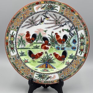 May include: A decorative ceramic plate featuring a detailed rooster design. The plate has a white background with a green and yellow landscape, and a colorful border. The roosters are red and black, and the plate is supported by a black stand.