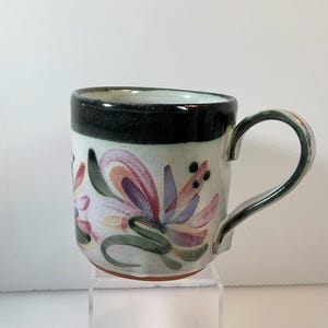 May include: Ceramic mug with a floral design. The mug features a black rim and handle, with a light green body. The floral pattern includes pink, purple, and green accents. The mug is approximately 4 inches tall.