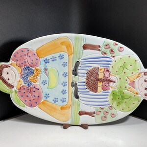 May include: Oval ceramic serving platter with a colorful, whimsical design. The platter features two stylized figures, one with a green head covering and the other with a pink head covering, and a central section with a basket and trees.