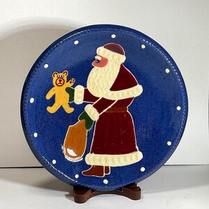 May include: A round, blue ceramic plate featuring a Santa Claus design. Santa is depicted in a red coat with white trim, holding a sack and a teddy bear. White dots encircle the plate's edge, adding a festive touch.