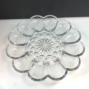 May include: Clear glass deviled egg serving platter with a scalloped edge. The center of the platter has a starburst design, and the outer edge has 12 individual egg-shaped compartments. This vintage serving dish is perfect for entertaining.