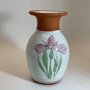 Puede incluir: Un jarr&oacute;n de cer&aacute;mica blanca con un borde marr&oacute;n y un dise&ntilde;o pintado de una flor de iris rosa con hojas verdes.