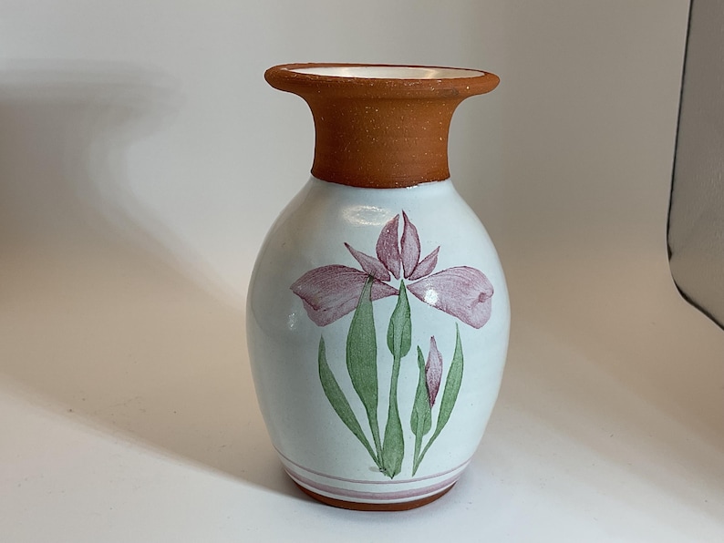 Puede incluir: Un jarr&oacute;n de cer&aacute;mica blanca con un borde marr&oacute;n y un dise&ntilde;o pintado de una flor de iris rosa con hojas verdes.