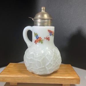 May include: White glass pitcher with a silver lid and handle. The pitcher features a textured, geometric pattern on the lower half and a painted floral design with blue, orange, and red berries. The pitcher is displayed on a wooden surface.