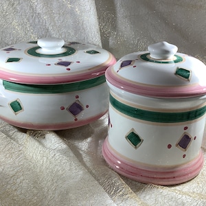 May include: Two ceramic canisters with lids. The canisters are white with pink, green, and purple geometric designs. The larger canister has a handle.