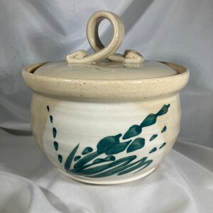 May include: A ceramic jar with a lid, featuring a cream-colored glaze and teal floral design. The lid has a unique handle. The jar is a round shape, suitable for kitchen or home decor.