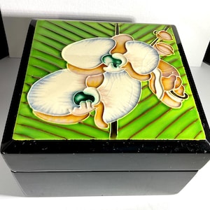 May include: A black wooden box with a green and white floral design on the lid. The design features a white orchid with green and orange accents on a green background with a diagonal striped pattern.