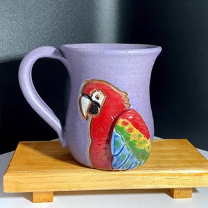 May include: A lavender ceramic mug with a sculpted, colorful parrot design. The parrot features red, yellow, green, and blue feathers. The mug has a curved handle and sits on a small wooden stand.