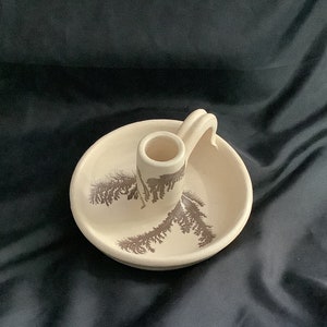 May include: A white ceramic candle holder with a brown tree design and a handle. The holder has a round base and a raised center for the candle.