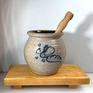 May include: A stoneware mortar and pestle set. The beige-colored mortar has a dark blue decorative design and a wooden pestle. The set is displayed on a small, light brown wooden stand.
