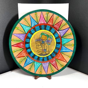 May include: A round, hand-painted decorative plate with a central figure and colorful geometric patterns. The plate features a green border, a tan background, and a variety of colors including blue, red, purple, and yellow. The plate is displayed on a small wooden stand.