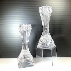 May include: Two clear crystal candle holders. The candle holders have a geometric design with vertical and angled lines. One is on a clear acrylic stand. The candle holders are designed to hold a single candle.