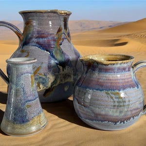 May include: Three ceramic pitchers in shades of blue, purple, and brown with a glossy finish. The pitchers are arranged on a sandy surface.