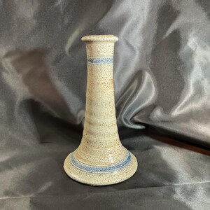 May include: A speckled beige ceramic candlestick holder with a flared base and a narrow neck. The piece features two horizontal bands of blue decorative detailing, one located near the top rim and another encircling the wide base of the object.