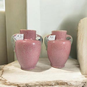May include: Two pink ceramic vases with handles and a textured surface. Each vase has a label with a number on it. The vases are displayed on a wooden surface.