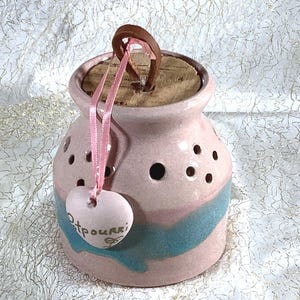 May include: A pink ceramic potpourri jar with a wooden lid and a pink ribbon. The jar has a blue and pink design and holes for air circulation. The jar has a heart-shaped tag that says "Potpourri".