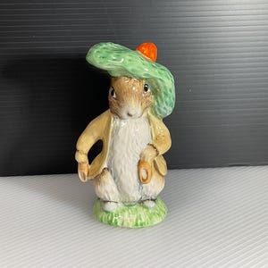 May include: A ceramic figurine of a rabbit wearing a green hat with an orange pompom, a tan jacket, and holding a small bag. The rabbit stands on a patch of green grass.