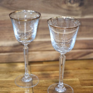 May include: Two clear glass stemmed drinking glasses with gold rims. The glasses are on a wooden surface.