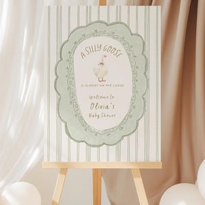Könnte beinhalten: Ein Schild für eine Babyparty auf einer hölzernen Staffelei. Das Schild zeigt eine skurrile Illustration einer Gans in einem dekorativen Rahmen. Der Text lautet: "A Silly Goose is Almost on the Loose. Welcome to Olivia's Baby Shower."