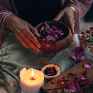 May include: A wooden bowl filled with dried rose petals and herbs, a lit candle, and a small bowl of dried herbs on a table covered in a green cloth.