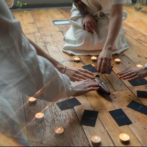 May include: Three people are gathered around a wooden table with candles and cards. They are performing a ritual or ceremony. The image is lit by candlelight, creating a warm and inviting atmosphere.
